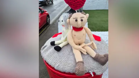 Susan Reddan A crocheted naked man and woman sitting on top of a post box with a knitted red balloon above