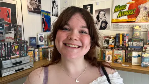 Grace is looking at the camera, she has dark hair and is wearing a white top and a crescent moon necklace.