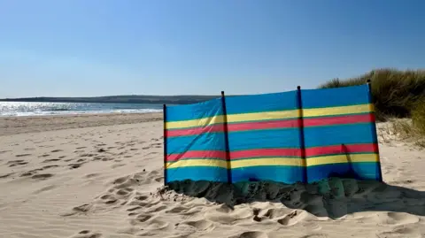 A sandy beach scene on a clear sunny day. A windbreak has been placed on the sand but there are no people in sight.