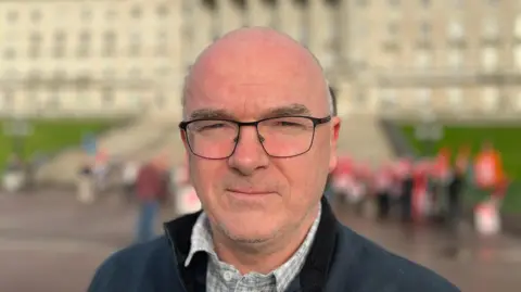 A bald man with black glasses and clear frames. He is wearing a navy sweatshirt and a grey shirt. The background is blurred but a white building and feint pictures of protesters can be pictured standing behind him