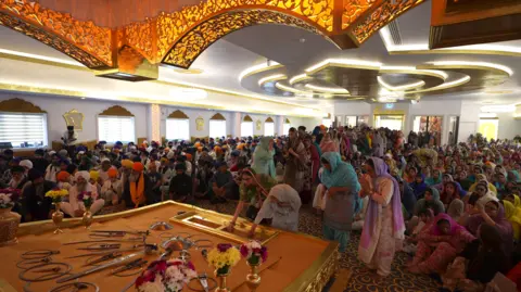Guru Nanak Sikh Gurdwara Men and woman sit on the floor on either side of the temple as a ceremony is held inside. Women in traditional dress place money on a tray in the centre of the room. An ornate gold decoration can be seen on the ceiling above a central, flat platform which has ceremonial knives on.