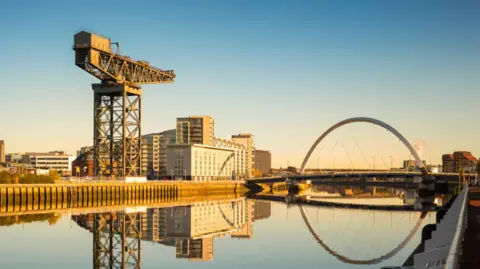 A view of the River Clyde in Glasgow with the Clyde Arc bridge and Finnieston Crane on the riverside. Blocks of flats and hotels line the water.