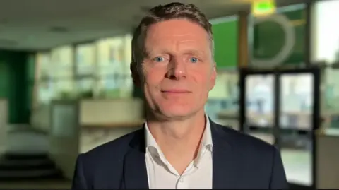 A close up of James in a hospital setting - blurred in the background. He is smiling to camera and wearing an open-necked white shirt and black jacket