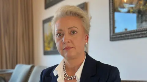 Magda is seated at a wooden table indoors, facing the camera. She is wearing a dark blazer over a light shirt. On the table are a laptop, papers, and a phone stand. The background includes framed artwork on the wall, upholstered chairs, window light, and hanging pendant lights.