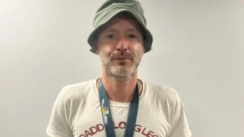 Philip Wallace stands looking directly into the camera. He is wearing a green bucket style hat, white t shirt and a lanyard.