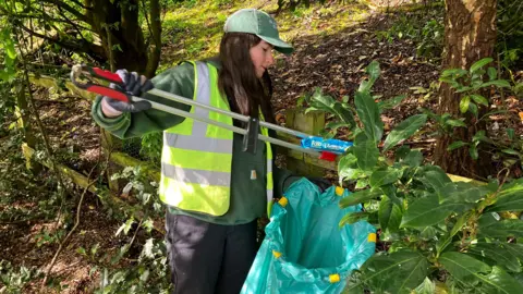 Amelia Prior in the process of clearing a piece of litter with her picker and a green trash bag.