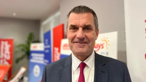 A man with short dark hair, greying at the temples, in a navy suit, with a white shirt and red tie.