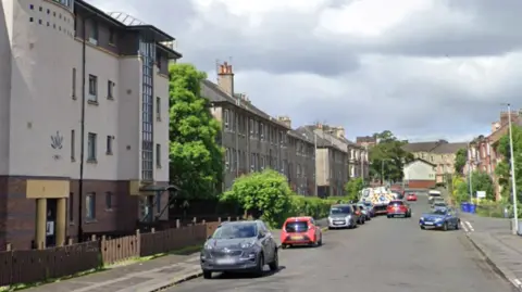Google Maps A residential street with flats and houses on either side. Cars are parked along both sides of the road and there is a some grass with trees and bushes on the right hand side of the road.