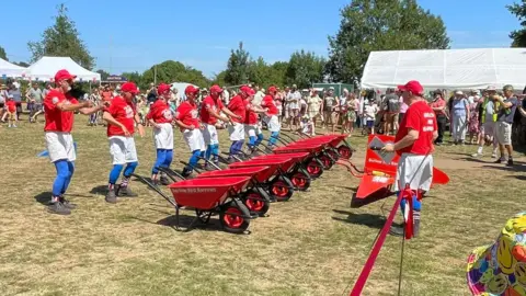 Barlow Red Barrows Eight people stood in a line behind a row of red wheelbarrows. They are dressed in red tops, white shorts and blue bottoms on their lower legs. A circle of people watches on.
