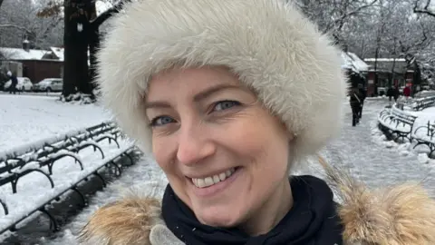 Christopher Jones Paula is standing in a park which has snow settled on the grass and pathway. She is wearing a fluffy white hat, a black jumper and a coat with a brown fluffy collar. 