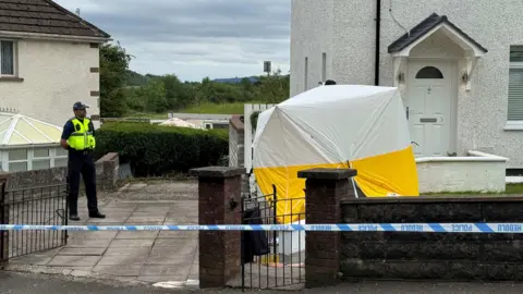 BBC Wales The scene at Rocky Road, Merthyr Tydfil. There's a forensic tent outside a white house with a brown brick wall. An officer is stood on the left hand side and there's police tape in front of the house. 