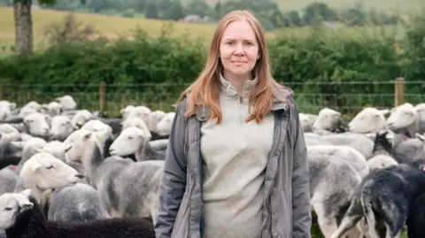 Joanne Coates Farmer's wife Helen Rebanks stands in a green grass field with many sheep behind her. The sheep are black and grey with white faces. Helen wears a beige fleece and a grey coat which is zipped open. She has auburn hair.