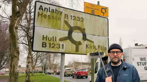 A man on the side of a road holding a large brush, of the type used by window cleaners, with a large, white, dirty road sign behind him. He is wearing a blue jacket and beanie hat. A white van is parked by the side of the road. On the back it says, "we clean stuff really good!" The road sign is covered in green moss. It has a roundabout symbol on it, with exits pointing to Anlaby, Hessle, B1232, Hull B1231 and Kirk Ella.