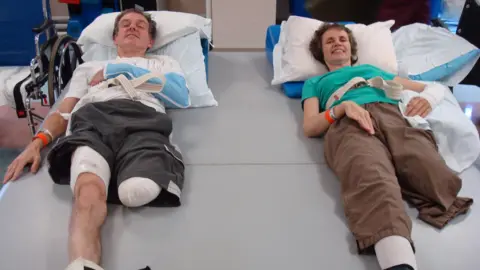 Chris and Denise Arthey A man and woman lying side by side on a hospital bed both with one leg amputated. 