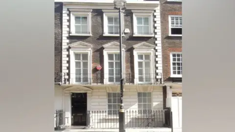 Front view of a 18th Century brick townhouse in London. It has a black front door and appears to be three storeys tall. There is a traditional black lamppost on the pavement outside