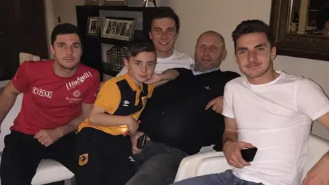 Family Handout The four brothers sat on the sofa with their late dad Chris, they all look young and are smiling at the camera. Tommy is on the far left of the picture, wearing a red boxing T-shirt, Rocco is wearing an orange and black Hull City tracksuit, with Joe in the background. Lewie, wearing a white T-shirt, is on the far right of the picture.