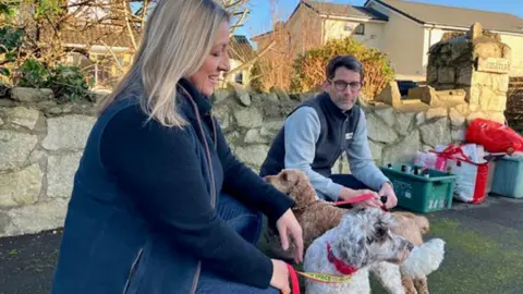 Jen, with blonde hair and wearing blue jacket, and Stuart, with glasses and blue body warmer, sitting with pets Margot (aged 5) and Willow (aged 4). They are outside near stone wall and bins. 