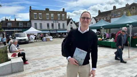 Ian Sollom Ian stares at the camera, holding a book which says St Neots Past. He wears a black top with beige trousers. He stands in St Neots Market Square. A number of stalls are in the background and there are people walking and sitting nearby.