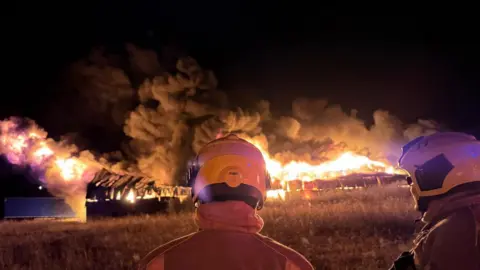 Two firefighters stood watching a large blaze which is lighting up the night sky in red and orange. There are huge plumes of smoke billowing from it 