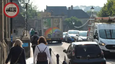 Bradford-on-Avon Town Council Two women, one blonde and one brunette walking with their backs to the camera towards a narrow pavement beside a bridge. Ahead of them a line of walkers in single file on the bridge. On the road on the bridge cars and vans queuing both sides.