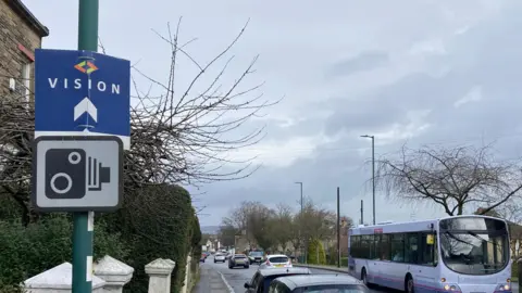 A speed camera roadsign, pictured against the backdrop of a busy road. A bus is on the opposite of the road.
