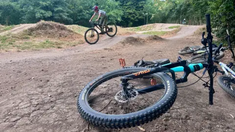 A cyclist wearing a helmet rides a bike over a dirt track surrounded by trees. In the foreground, two bikes lie on the ground beside the trail, with one wheel prominently visible front and centre.