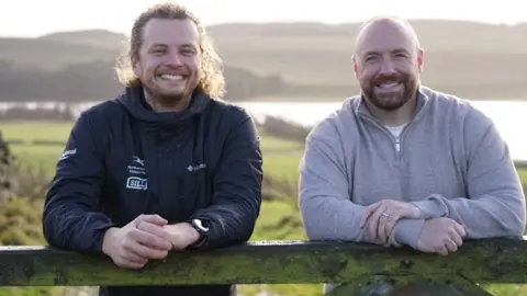 Adam Ormston/NNPA Jonny Snape, nature recovery ranger, and Matthew Squires, from The Squires Foundation. They are outdoors in an area of open countryside, with a lake in the background. They are both leaning on a gate or fence and smiling at the camera. Mr Snape is wearing a dark outdoor jacket, he has long blond curly hair tied back and a short beard. Mr Squires is bald with a trimmed brown beard. He is wearing a grey jumper.