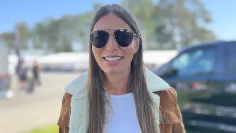Jessica has long, straight light brown hair with a brown cord jacket with a white fluffy colour. She is smiling and wearing aviator sunglasses, on a sunny day.