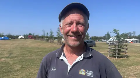 Jules smiles at the camera, standing in the middle of a camping field with caravans and tents parked around it. He has short, greying hair and a beard, and wears a flat cap and a navy blue polo shirt with the campsite branding and his name on it. 