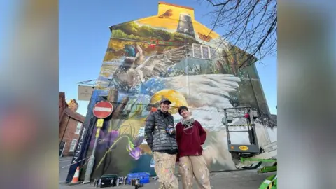 Two people wearing baggy clothes covered in paint specks smiling and standing in front of a mural they have created on the side of a dental practice. The mural depicts the River Slea with a canal boat and mill, and in the foreground is a duck and a swan splashing their wings in the water.