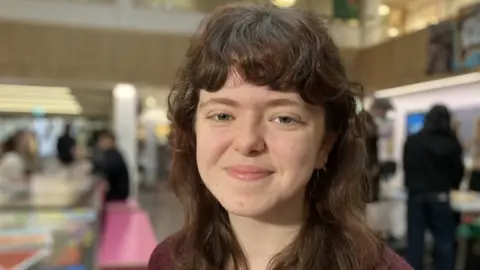 Alex Dunlop/BBC A young woman with long auburn hair and a full fringe smiles at the camera.