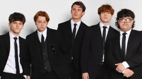 The five members of Swindled are standing in a line and are all wearing black suits, black ties and white shirts