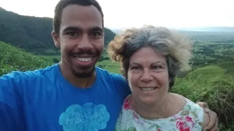 Will and Jane Plastow stand next to each other. Will has his arm around Jane. They are on a hill, with a view of a green valley behind them.