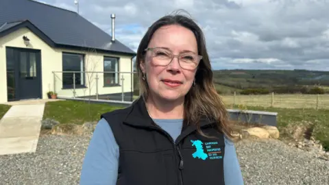 Tania Dutnell, estate agent. Tania has long brown hair reaching to her shoulders and has glasses and silver dangling earrings. She is wearing a light blue long sleeved top with a black bodywarmer on top. The bodywarmer has a logo with a light blue map of Wales and the words 'Cardigan Bay properties'. Behind her is a cream house with black doors and fields behind. 