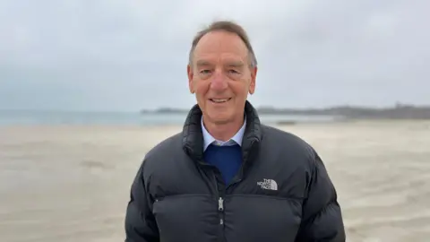 BBC A man wearing a black puffa jacket, standing on a beach