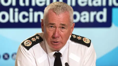 Police Service of Northern Ireland Chief Constable Jon Boutcher is speaking into a microphone. He is sitting in front of a large blue poster which has the words 'Northern Ireland Policing Board' on it. He is wearing a white shirt which has police badges on both shoulders and also a black tie. 