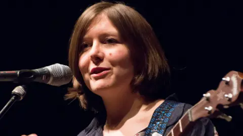 Getty Images Emily Portman holding a guitar and speaking into a microphone on stage