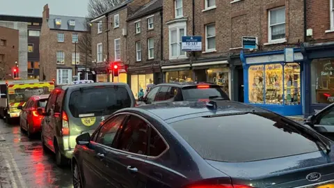 Traffic congestion on a busy road, a line of cars and service vehicles wait at a red traffic light junction.