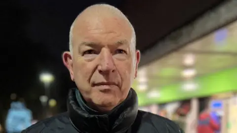 Jim Cunningham has cropped grey hair and is wearing a thick black coat. The background is blurred. It is dark.