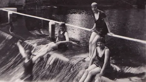 BPEOSA collection A black and white image of four women in water, by a metal bar. They all have swimming costumes on and hats, one woman is slightly blurred as she is sliding down a bank. One woman is standing and two are sat down. 