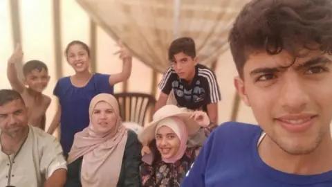 al-Dalou family photo Selfie photo taken by Sha'aban al-Dalou of him and his family. He is a 19-year-old man wearing a blue T-shirt, standing in the front, with six relatives from childhood to middle age smiling behind him. Some children are doing a thumbs up gesture.