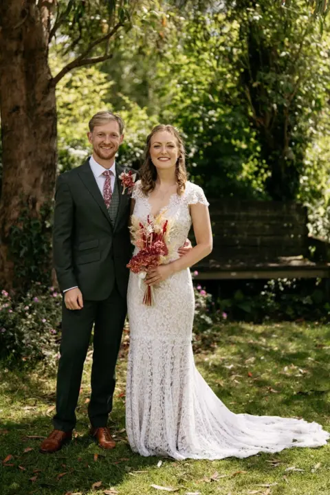 Dan Wimble Weddings A young couple pose for a photo on their wedding day. They are outdoors, surrounded by trees and greenery. The groom wears a dark suit with tan shoes and the bride wears a deep v-neck, white lace, fishtail wedding dress. 
