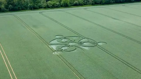StillPho ToMalone View from above of a crop circle in a green crop field. Three eye-shapes with more circles inside them and three lines each with their own circles going through the middle of them
