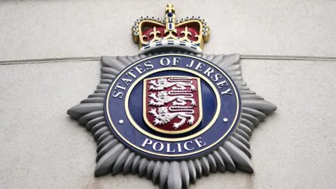 The States of Jersey Police crest mounted on a wall. It is shaped like a septagram within which is a circle holding the Jersey shield featuring three lions and the words STATES OF JERSEY POLICE around the outside. There is a crown at the top of the crest.
