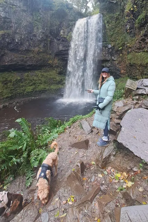 Leah Matthews A woman with long bropwn hair wearing a long, light blue blue puffer coat, a black cap and blue walking boots. She is stood in front of a waterfall and surrounded by cliffs. In the forefront of the picture are two small, fluffy brown dogs wearing harnesses.