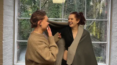 Donna Smith Two girls looking at each other laughing. The girl on the left is wearing a beige jumper and has her hand up by her mouth. She has a tattoo on the back of her neck. The girl on the right is wearing a black jumper and has a grey blanket wrapped around her. 
