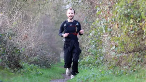 Stuart Woodward/BBC Jack runs towards the camera along a countryside footpath which has hedges on either side. He wears white trainers and dark navy running top and trousers.
