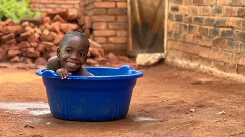 Rachael sitting in a blue plastic tub, presumably used for washing. You can see her smiling face over the edge of the tub, which is sitting in the sandy backyard of a house.
