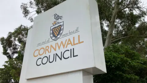 BBC Cornwall Council sign in front of some trees. The sign is white and square with yellow and black writing. 