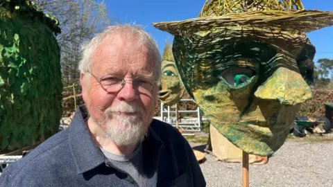 BBC/Andy Watson John Parkinson wearing a blue over shirt with a grey T-shirt, stands in front of a green puppet head which he is constructing for May Day. 
